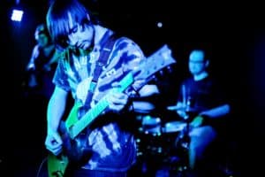 Two band members playing together on stage, with one guitarist in the foreground and the second musician partially visible behind.