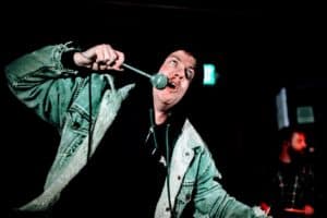 A performer passionately singing into the microphone while gripping a large spoon like a prop during a live concert.