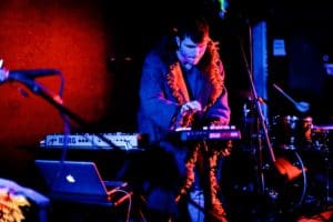 Musician performing live on stage, surrounded by electronic instruments and a laptop, creating an atmospheric and immersive setup against a vibrant red background.