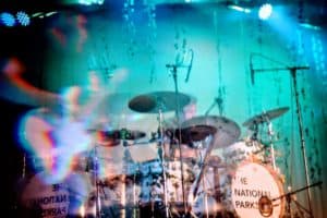 A blurred, dynamic stage shot of a drummer from The National Parks, with vibrant blue and teal lighting creating a ghostly double-exposure effect.