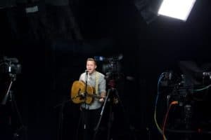 Artist playing an acoustic guitar in a professional studio setting, flanked by multiple cameras and lighting equipment, preparing for a video recording session.