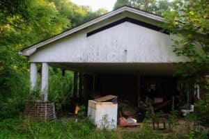 A rundown carport with a sagging, white-painted roof. The area is littered with old furniture, debris, and a rusty refrigerator. Wild vegetation encroaches on the structure, creating an eerie scene of neglect.