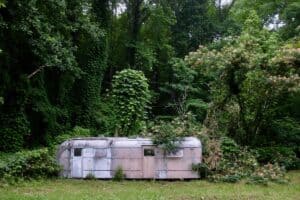 An old, weathered trailer sits abandoned in a clearing, partially overtaken by nature. Vines and plants creep over the faded pink exterior, blending it into the dense forest surroundings. The trailer's worn, dilapidated state reflects a forgotten chapter of the past.